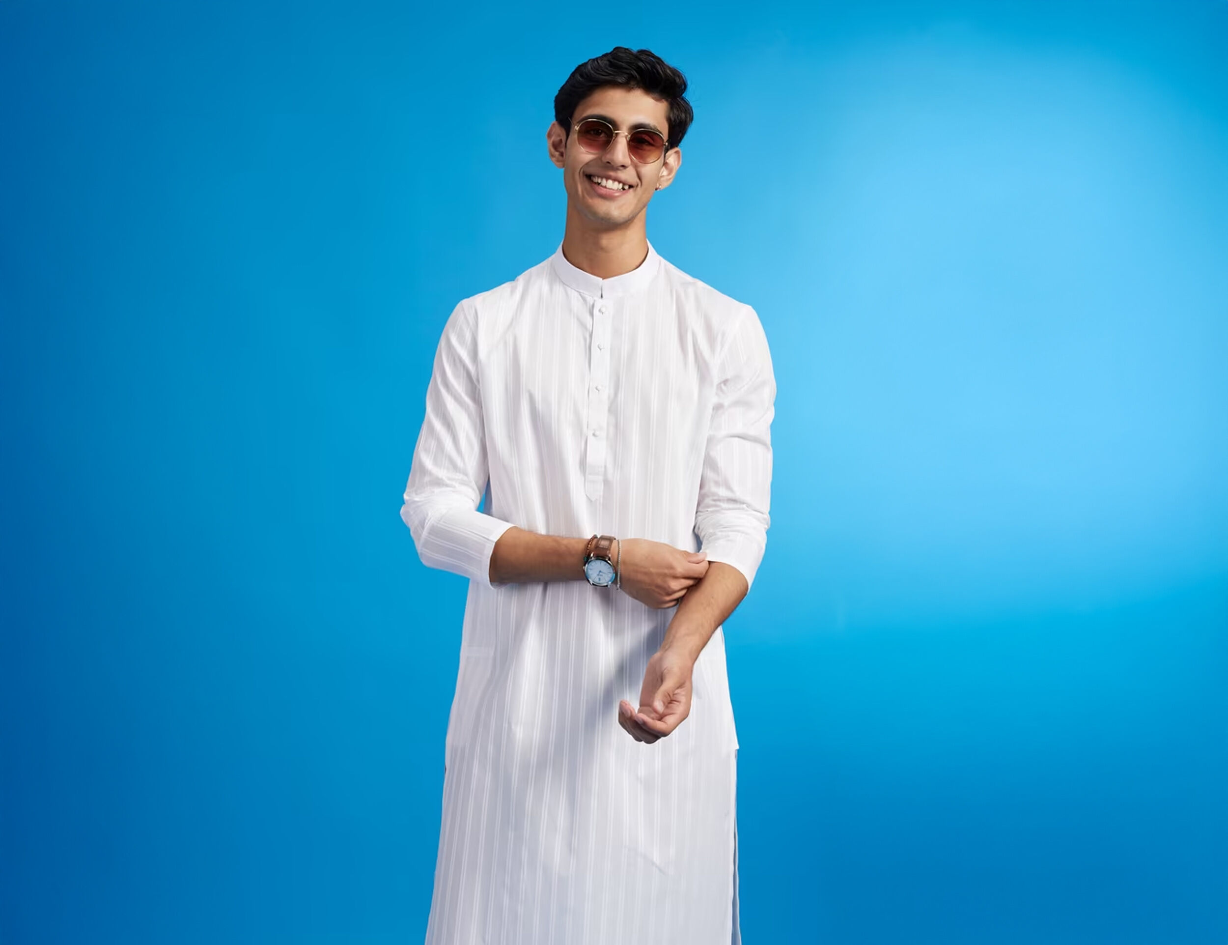 Smiling man wearing a white chikankari kurta and sunglasses, adjusting his sleeve while posing confidently against a bright blue background, blending traditional elegance with modern flair.